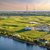 Großes Golfplatzareal mit grünen Rasenflächen und Wasserlauf im Vordergrund, im Hintergrund Industrieanlagen mit hohen Schornsteinen und Windrädern bei Sonnenuntergang.