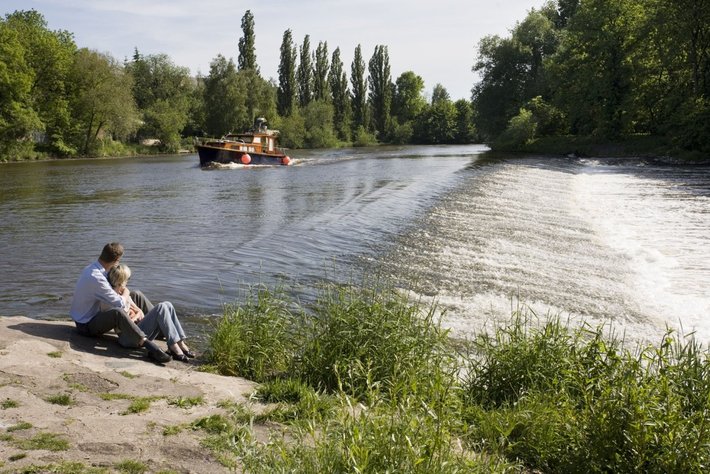 Eine Person sitzt am Ufer eines Flusses und??éct einen Vorbeifahrenden Bootstraktor mit Anhänger.

Normalerweise gemütliche Naturansicht am Gewässer mit fließendem Wasser und background Gründ???igkeit.