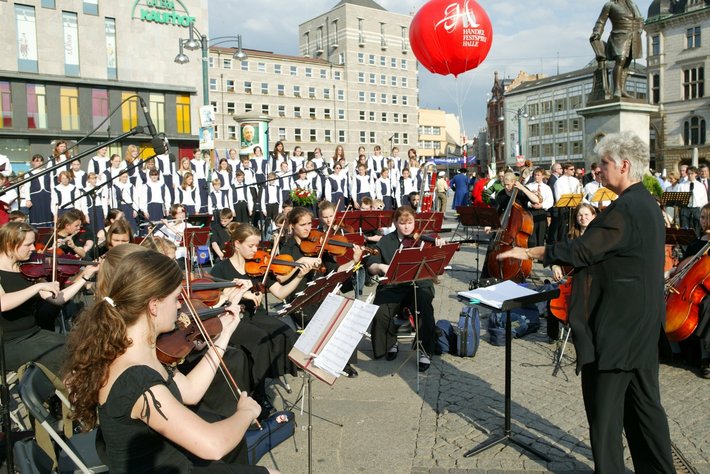 Große Orchesterprobe im Freien mit vielen Musikern, die Streichinstrumente wie Violinen und Celli spielen. Ein Dirigent steht vor den Musikern und leitet das Orchester.