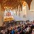 Großer Kirchenraum mit Empore, auf der ein Orchester mit zahlreichen Musikern sitzt und ein Konzert gibt. Im Saal sitzen viele Zuschauer auf Holzbänken und verfolgen die Aufführung.