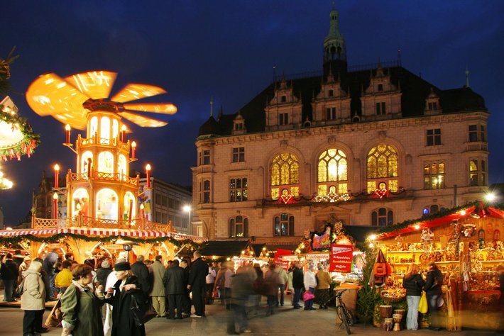 Ein belebter Weihnachtsmarkt bei Nacht mitembleuteter Architektur, zuz merklich historischen Bauwerken. Menschen schlendern zwischen Ständen mit festlicher Dekoration. Girlanden schmücken die Szene und eine beleuchtete große Holzkrippe ziert den Mittelpunkt.
