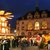 Ein belebter Weihnachtsmarkt bei Nacht mitembleuteter Architektur, zuz merklich historischen Bauwerken. Menschen schlendern zwischen Ständen mit festlicher Dekoration. Girlanden schmücken die Szene und eine beleuchtete große Holzkrippe ziert den Mittelpunkt.