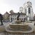 Bronzestatue eines springenden Pferdes mit Reiter im Zentrum eines runden Brunnenbeckens, umgeben von vier weiteren Reiterstatuen auf Sockeln, vor historischer Kirche mit Turm und klassizistischer Fassade.