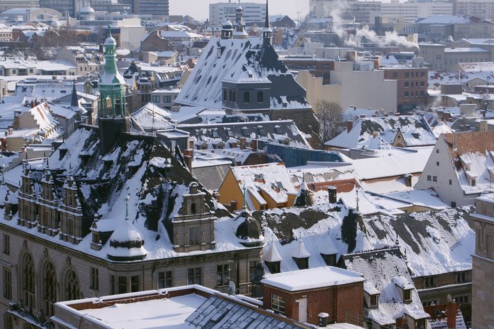 Ein Bild einer verschneiten StadtAvec historischen und klassischen Gebäuden, wobei besonders eine charaktervolle Kirche mit grünem Turm und spitzem Dach im Vordergrund sichtbar ist.