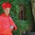 Ein älterer Mann posiert mit einem roten byzantinischen Mantel und einer roten Bischofsmütze vor einer grün bewachsenen Mauer.
Er scheint lächelnd und trägt die traditionelle katholische liturgische Gewandung.