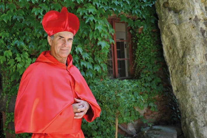 Ein älterer Mann posiert mit einem roten byzantinischen Mantel und einer roten Bischofsmütze vor einer grün bewachsenen Mauer.
Er scheint lächelnd und trägt die traditionelle katholische liturgische Gewandung.
