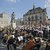 Großer Chor und Orchester mit zahlreichen Musikern und Sängerinnen, die auf einem öffentlichen Platz vor historischer Architektur und einem Denkmal mit Reiterstatue gemeinsam musizieren.