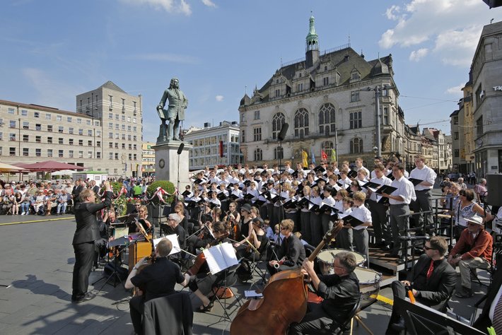 Großer Chor und Orchester mit zahlreichen Musikern und Sängerinnen, die auf einem öffentlichen Platz vor historischer Architektur und einem Denkmal mit Reiterstatue gemeinsam musizieren.