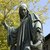 Stehende Bronzestatue einer Person mit langem Gewand und Kapuze, die mit einer Hand nach vorne deutet, während die andere Hand ein Buch oder Schriftrolle hält. Im Hintergrund ein Baum mit grünen Blättern.