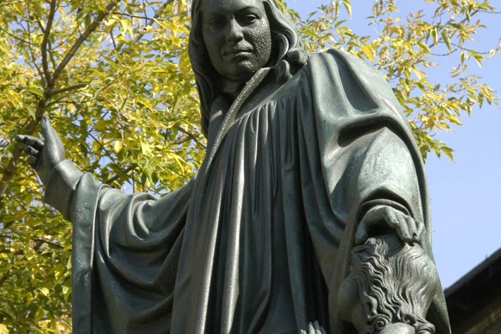 Stehende Bronzestatue einer Person mit langem Gewand und Kapuze, die mit einer Hand nach vorne deutet, während die andere Hand ein Buch oder Schriftrolle hält. Im Hintergrund ein Baum mit grünen Blättern.