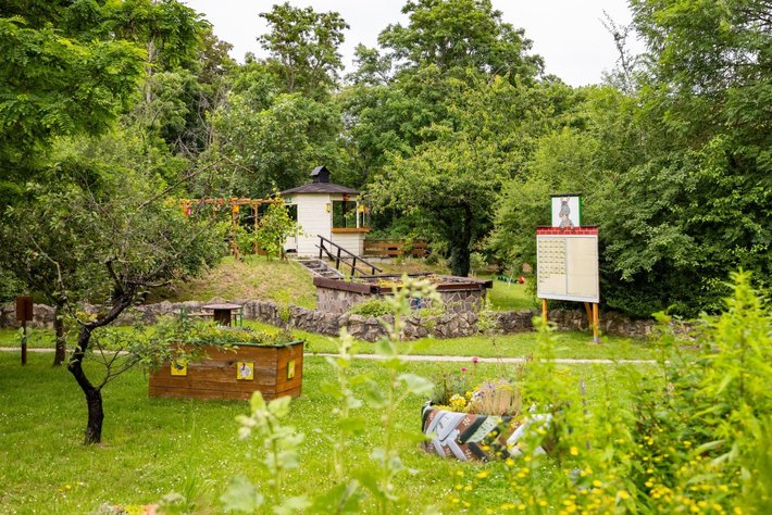 Idyllisches Gartenhaus mit Holztreppe und umliegendem Garten, umgeben von Bäumen und Sträuchern. Im Vordergrund ein Hochbeet mit Kräutern und ein Infoschild mit rotem Rahmen.