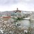 Ein winterliches Stadtpanorama mit Schnee bedeckt: Eine Brücke überspannt einen Fluss, im Hintergrund erhebt sich eine festungsartige, stilisierte Burg auf einem Hügel. Bäume und Gebäude sind mit einer Schneeschicht überzogen.
