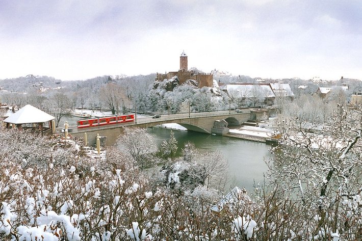 Ein winterliches Stadtpanorama mit Schnee bedeckt: Eine Brücke überspannt einen Fluss, im Hintergrund erhebt sich eine festungsartige, stilisierte Burg auf einem Hügel. Bäume und Gebäude sind mit einer Schneeschicht überzogen.