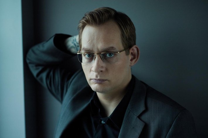 Ein Mann mit kurz geschnittenen Haaren, Brille und ernstem Blick, der ein dunkles Sakko trägt und sich leicht nach hinten lehnt.
