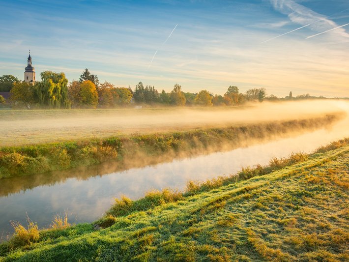 Frühnebel über einem ruhigen Fluss in einer Landschaft mit grünen Wiesen und goldgelb leuchtenden Bäumen im Herbst. Im Hintergrund sind sanfte Hügel und eine Silhouette von Bäumen sowie ein Kirchturm zu erkennen.