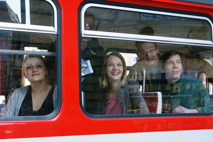 Drei Personen sitzen lächelnd in einem roten Zugabteil, schauen aus dem Fenster und wirken entspannt. Eine Frau links, eine junge Frau in der Mitte und ein älterer Herr mit Brille rechts.