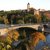 Eine historische Burg auf einem Hügel mit mittelalterlichem Turm, im Hintergrund. Davor führt eine Brücke über einen Fluss, auf der Fahrzeuge und ein Boot fahren. Im Herbst bunte Bäume säumen die Ufer.