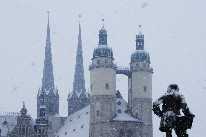 In dieser winterlichen Szene sind Gebäude mit typischen gotischen Spitzen und Türmchen im Schneegestöber zu sehen. Davor steht eine Person zu zweit eng aneinandergedrückt, sichtbar durch Umhänge bekleidet. Kälte und Romantik prägen den Frostsnow-Spaziergang unter disse Ornamenten.