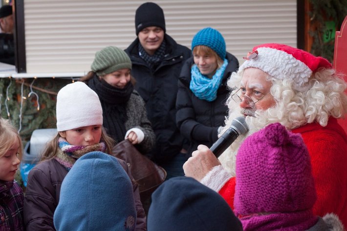 Weihnachtsmann mit rotem Mantel und weißem Bart hält ein Mikrofon und spricht zu einer Gruppe winterlich bekleideter Kinder und Erwachsener, die gespannt zuhören.