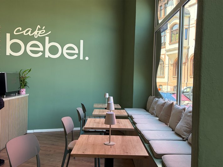 Moderner Café-Bereich mit hellen Holztischen und Stühlen, grünen Wänden und großen Fenstern, die Tageslicht hereinlassen. Im Hintergrund ein kleiner Arbeitsplatz mit Computer.