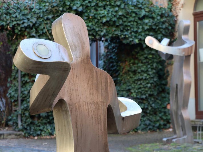 Holzskulptur eines sitzenden Menschen mit großen, runden Ohrmuscheln und geschlossenen Augen, die eine meditative oder nachdenkliche Haltung ausstrahlt.