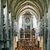 Großer Chorraum mit steinernen Bögen und Säulen, in dem ein Chor mit vielen Sängern und Instrumenten auf der Bühne sitzt. Im Hintergrund prächtiger Altar mit Goldverzierungen, Gemälden und Kerzen.