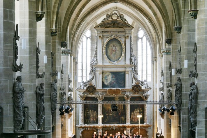 Großer Chorraum mit steinernen Bögen und Säulen, in dem ein Chor mit vielen Sängern und Instrumenten auf der Bühne sitzt. Im Hintergrund prächtiger Altar mit Goldverzierungen, Gemälden und Kerzen.