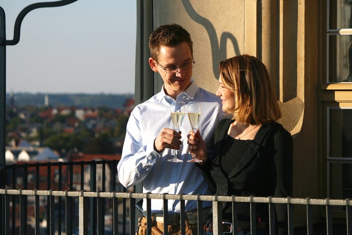 Zwei Personen stehen auf einem Balkon und stoßen mit Sektgläsern an, lächelnd und sich gegenüber. Im Hintergrund ist eine Stadtlandschaft mit Gebäuden und Himmel zu sehen.