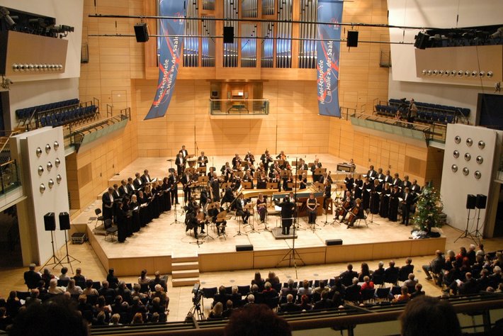 Ein großes Orchester mit zahlreichen Musikern spielt auf einer Bühne vor einem Publikum. Im Hintergrund sind zwei große Banner mit blauen und roten Mustern zu sehen. Die Szene spielt in einem Konzertsaal mit guter Akustik.