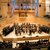 Ein großes Orchester mit zahlreichen Musikern spielt auf einer Bühne vor einem Publikum. Im Hintergrund sind zwei große Banner mit blauen und roten Mustern zu sehen. Die Szene spielt in einem Konzertsaal mit guter Akustik.