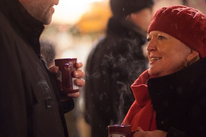 Zwei Personen unterhalten sich bei kaltem Wetter und halten je eine warme Tasse.

Die Person auf der linken Seite trägt eine dunkle Mütze und pelzbesetzten Umhang, die Person rechts einen roten Schal und eine rote Mütze.