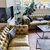 Gemütlicher Wohnraum mit zwei großen, cremefarbenen Ledersofas mit Kissen, einem kleinen Couchtisch in der Mitte und einem weiteren Tisch mit Pflanzen und Getränken. Im Hintergrund ein Kamin und große Fenster mit Blick auf eine Straße.