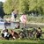 Die Aufnahme zeigt eine Gruppe Menschen in der Nähe eines ruhigen Flusses, die sich auf einer Wiese aufhalten. Vier Personen sitzen oder knien auf dem Gras, dazu stehen mehrere Fahrräder daneben. Kinder und Erwachsene wirken entspannt, dahinter sind aufgezeltete Boote und eine Banntafel zu sehen.