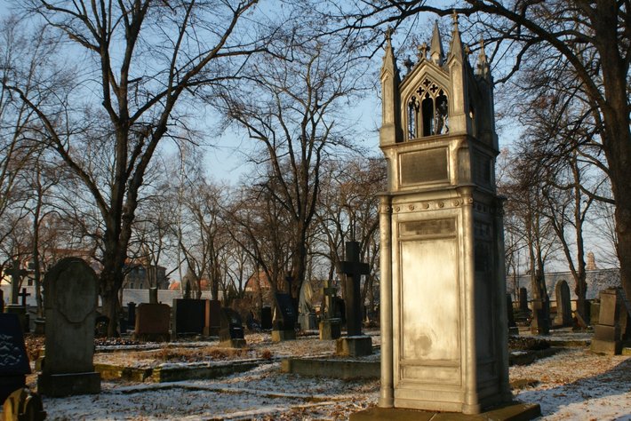 Alternativtext für sehbehinderte Personen:

Einfriedeter Friedhof auf einer leicht schneebedeckten Fläche. Im Vordergrund steht ein einzelnes, altmodisches Grabmal aus Stein mit ornamentalen Details undURSSchrifttafel, umgeben von alten Bäumen ohne Blätter und anderen Gräbern einheitlich lindgräulich und historisch gestaltet.