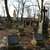 Ein ruhiger, überdachter Friedhof mit mehreren alten Grabsteinen unterschiedlichen Stils auf einem mit Laub bedeckten Rasenboden im Herbst. Im Hintergrund leere, schlanke Bäume und eine siluettierte Backsteinkirche.