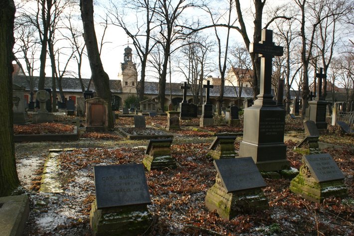 Ein ruhiger, überdachter Friedhof mit mehreren alten Grabsteinen unterschiedlichen Stils auf einem mit Laub bedeckten Rasenboden im Herbst. Im Hintergrund leere, schlanke Bäume und eine siluettierte Backsteinkirche.