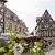 Historisches Fachwerkhaus mit typischer Giebelarchitektur in einer malerischen Altstadtgasse. Vor dem Gebäude befindet sich ein Außenbereich mit Blumenbeeten, weißen Blüten und Sonnenschirmen eines Cafés oder Restaurants. Zwei Personen gehen auf dem gepflasterten Platz.