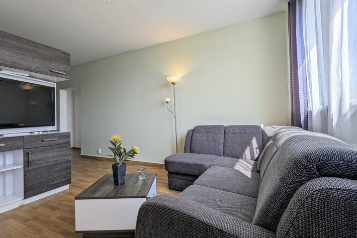 Gemütliches Wohnzimmer mit grauer Couchgarnitur, kleiner Glastischmitte, darauf eine Vase mit gelben Blumen, Fernseher in einer dunklen Einbauwand und einem Stehlampe mit warmem Licht.
