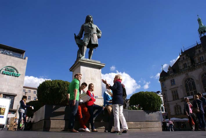 Éine Gruppe von vier erwachsenen Menschen steht vor einer Bronze Statue eines Mannes auf einem altasartigen Sockel an einem sonnigen Platz.
Die Statue scheint bedeutend oder historisch zu sein und hierfür bilden sie eine geschlossene Gruppe.