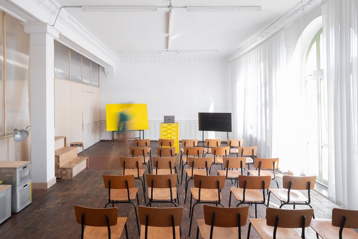 Leerer, hell erleuchteter Seminarraum mit vielen braunen Holzstühlen in Reihen, großer gelber Leinwand oder Wandfläche auf der linken Seite und einem Bildschirm auf der rechten Seite. Große Fenster mit Tageslicht an der Rückwand.