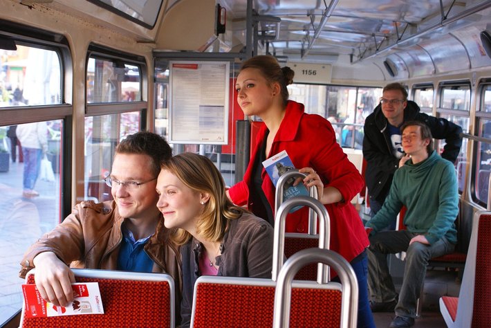 Ein Paar sitzt lächelnd nebeneinander im Bus, während eine Frau in rotem Mantel danebensteht und auf ihr Handy schaut. Im Hintergrund weitere Fahrgäste auf roten Sitzen.