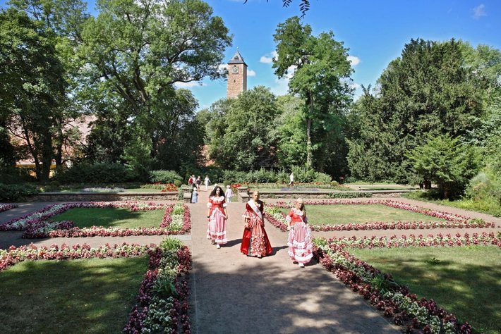 Drei Personen in traditioneller, rot-weiß karierter Kleidung lactate auf einem gepflegten Kiesweg durch einen blühenden Garten mit Üppigen, roten und weißen Blumenbeeten.