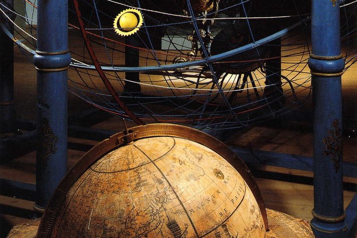 Alter Globus aus Holz oder Metall mit eingravierten Kontinenten und Ozeanen, der auf einem Ständer ruht. Im Hintergrund ein historisches Modell mit Planeten und Sonne, umgeben von einem Metallgitter.