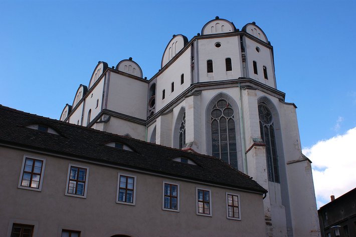 Gotische Kirche mit zwei markanten Rundtürmen, hohem Mittelschiff und großen, spitzbogigen Fenstern, darunter ein großes Rosettenfenster. Hell verputzte Fassade mit dunklem Dach.