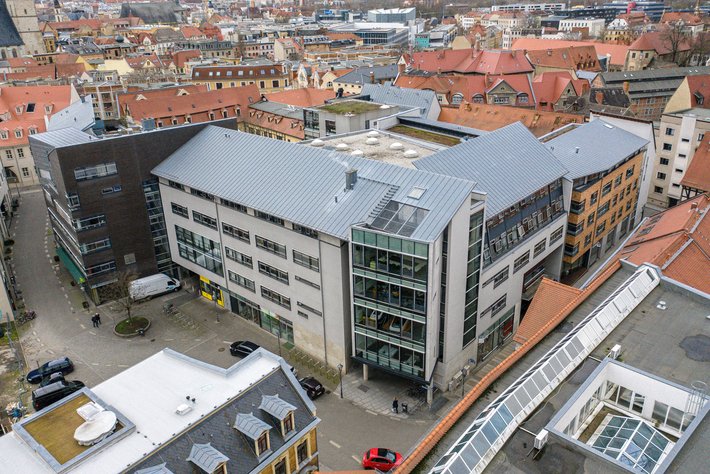 Moderne Bürogebäude mit hellen Fassaden und großen Glasflächen, umgeben von historischen roten Dachhäusern in einer Stadtlandschaft. Im Innenhof sind Fahrzeuge und ein kleiner Parkbereich zu erkennen.