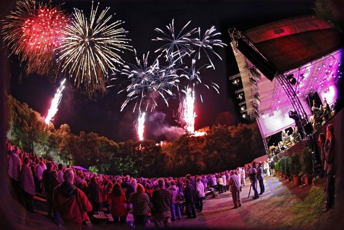 Große Menschenmenge bei Nacht, die ein spektakuläres Feuerwerk mit roten, weißen und goldenen Raketen bewundert, das am Himmel explodiert.
