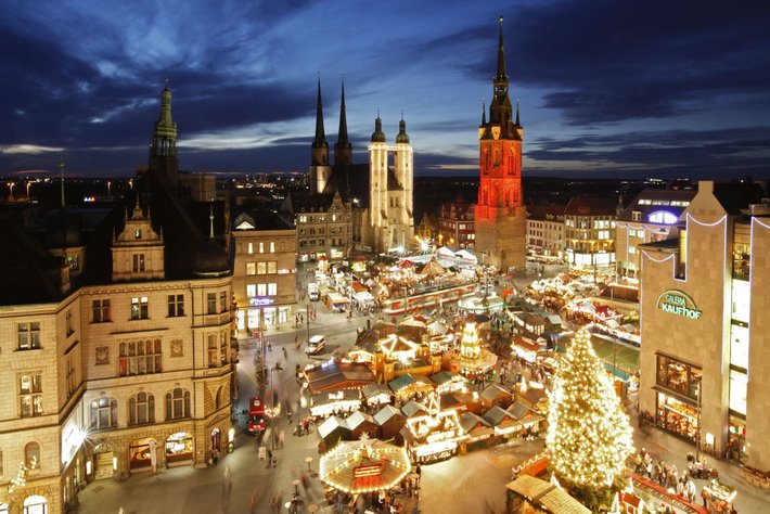 Diese Takoaufnahme zeigt einen festlich beleuchteten Weihnachtsmarkt auf einem städtischen Hauptplatz. Im Hintergrund ragen historische Kirchen und Gebäude hervor. Die Stände laden mit Dekorationen ein und ein großer Weihnachtsbaum erstrahlt in der Mitte. Die Szene ist sehr besinnlich und festlich.