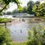 Eín idyllisches Flußufer mit mehreren Enten, die auf dem Wasser schwimmen. Häuserboote oder Fahrgastschiffe liegen an; Menschen Behören am Ufer. Grüne Umgebung mit Bäumen und Sträuchern. Ruhige Naturatmosphäre.