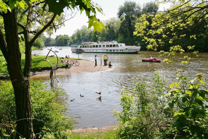 Eín idyllisches Flußufer mit mehreren Enten, die auf dem Wasser schwimmen. Häuserboote oder Fahrgastschiffe liegen an; Menschen Behören am Ufer. Grüne Umgebung mit Bäumen und Sträuchern. Ruhige Naturatmosphäre.