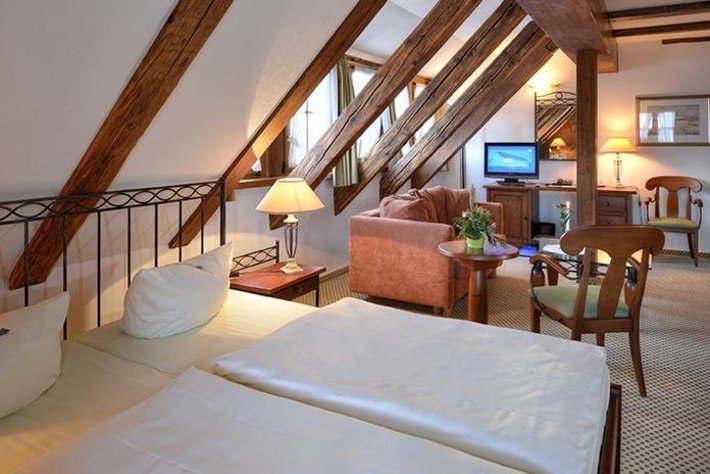 Dieses Bild zeigt ein gemütliches Zimmer unter einem Satteldach.
Im Raum befinden sich zwei Request-Betten mit weißer Bettwäsche.
Dazu gibt es einen angesagten Wohnbereich mit Couchtisch und Sofa, deskmit Pflanzen-Deko. Anschließend folgt ein TV-Bereich und ein Sessel mit Tisch.
Das Zimmer wirkt hell und einladend.